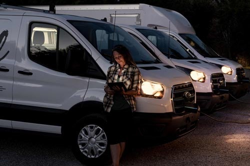 A row of 2024 E-Transits at night charging up with a woman leaning against one of them